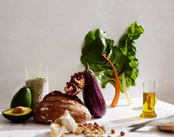 A still life of a collection of foods on a marble surface against a light gray wall. The foods include avocado, beans, garlic, eggplant, olive oil, pomegranate, nuts, whole-grain bread, fish and leafy greens.